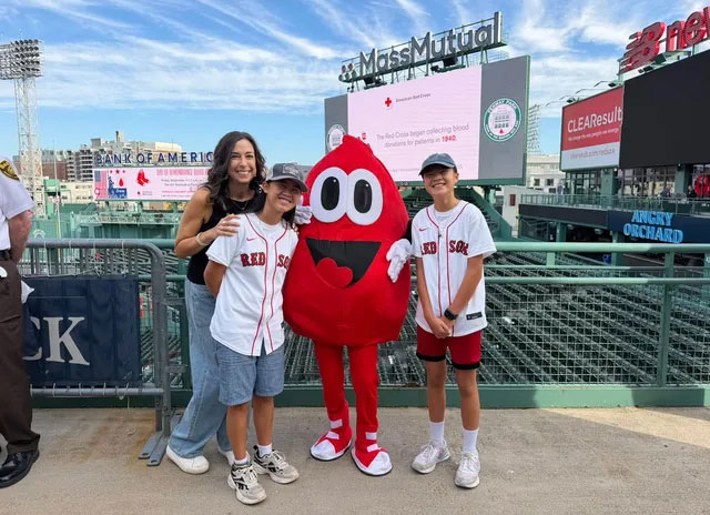 a woman and two kids with Red Sox shirts standing next to a blood drop mascot in Fenway park.