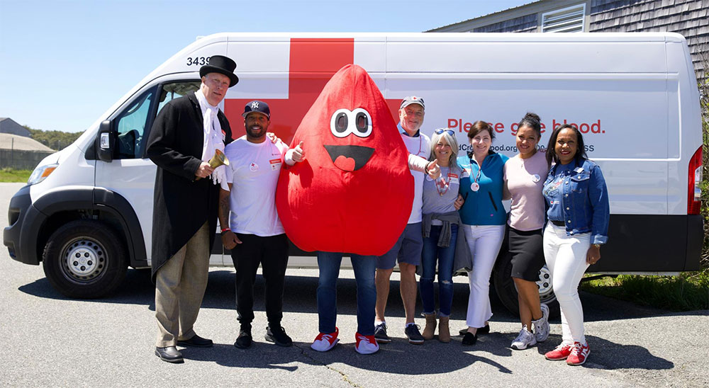 Group picture of blood donors, blood drop mascot, a guy with a top hat, coat and bell with a Red Cross van in the background.