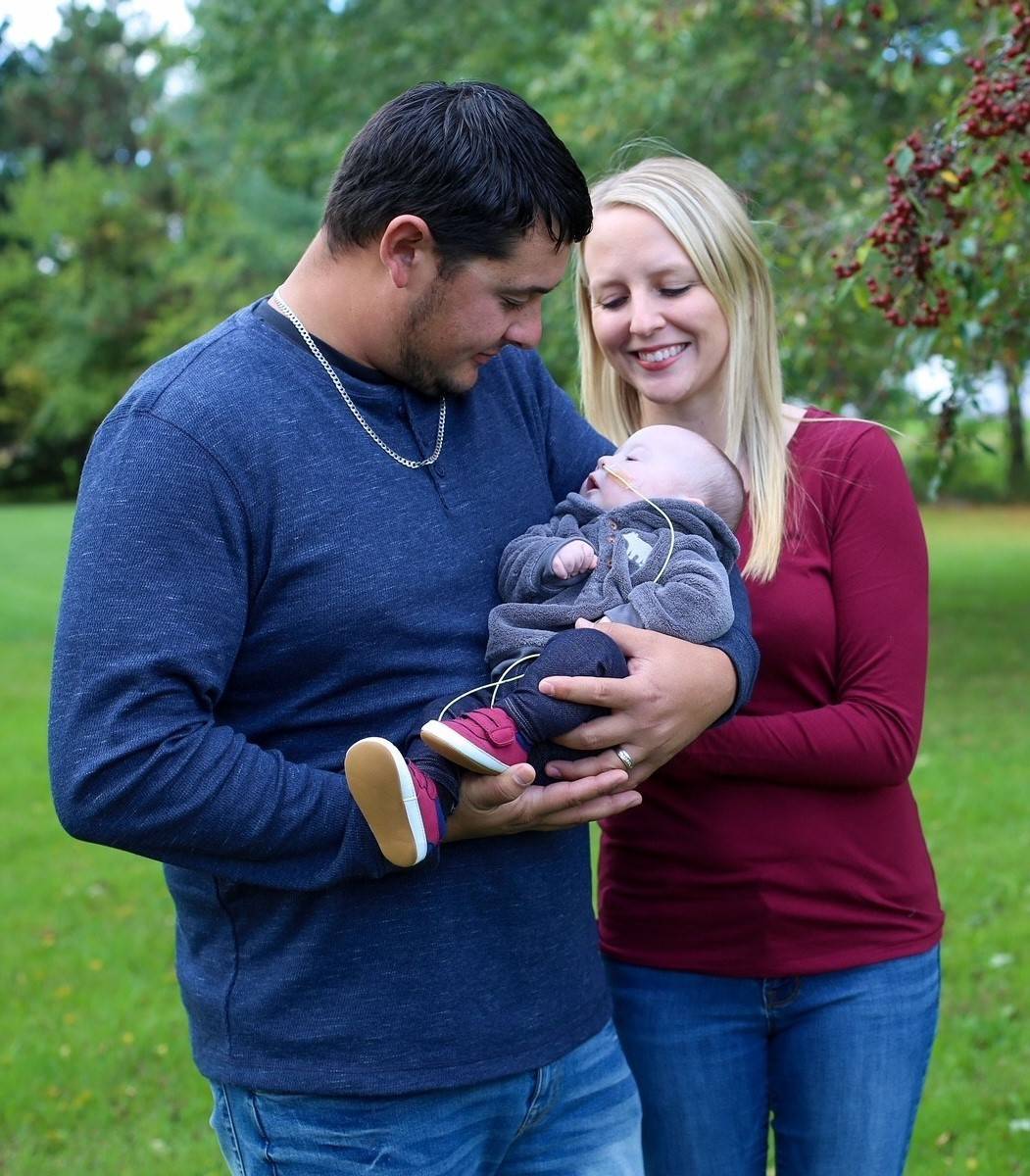 Parents outside holding baby and smiling. Baby has breathing tube in nose.