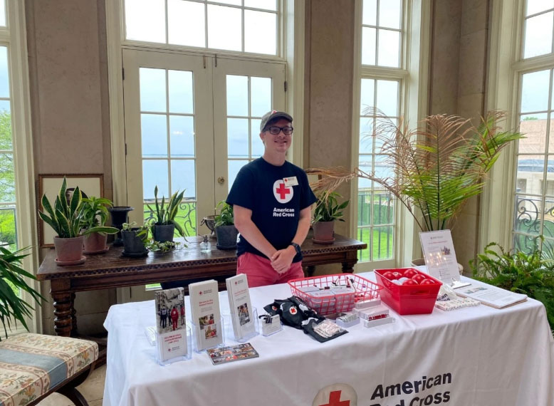Tyler White in Red Cross shirt standing behind a table full of Red Cross promotional items.