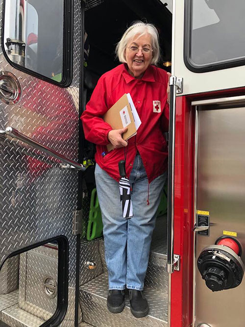 PJ Doyle wearing a Red Cross jacket and holding paperwork while stanging on the stairs of a firetruck .