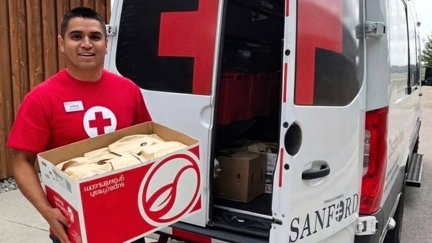 Richard Felix in a Red Cross shirt holds a box of supplies beside an open Red Cross van.