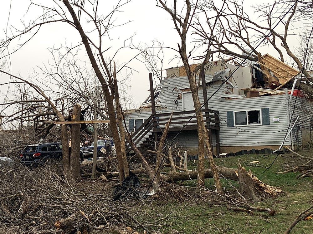 tornado damaged house with broken trees around it.