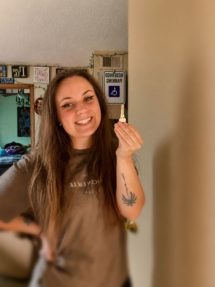 Hannah Vollintine smiling with long brown hair holds a house key. She wears a brown T-shirt and has a tattoo on her forearm. The background shows a reserved parking sign and home decor.