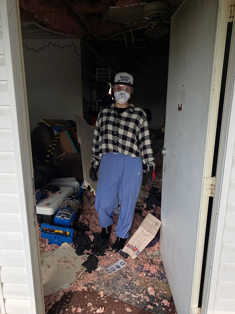 Hannah Vollintine dressed in casual clothing with a face mask stands in a cluttered, messy room with debris and insulation covering the floor.