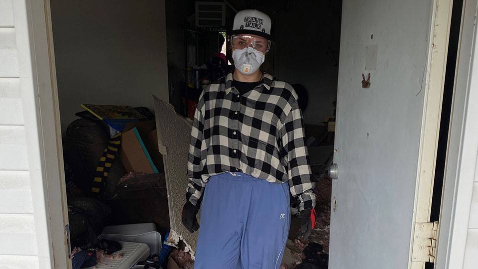 Hannah Vollintine dressed in casual clothing with a face mask stands in a cluttered, messy room with debris and insulation covering the floor.