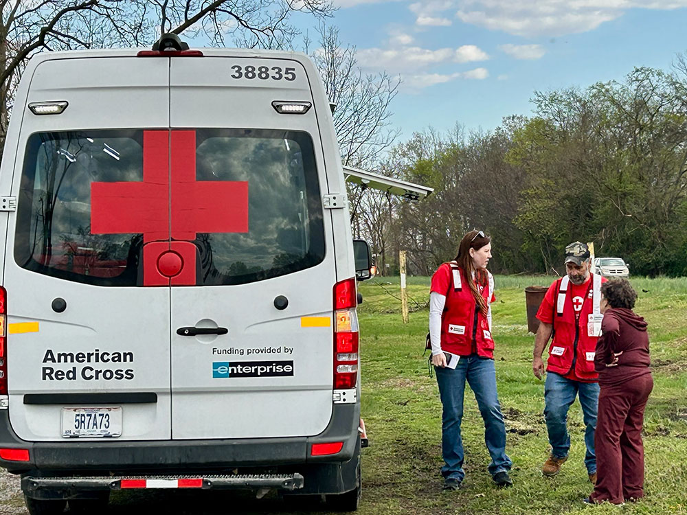 Red Cross vehicle parked on the side of the road with Red Cross volunteers speaking to a person.