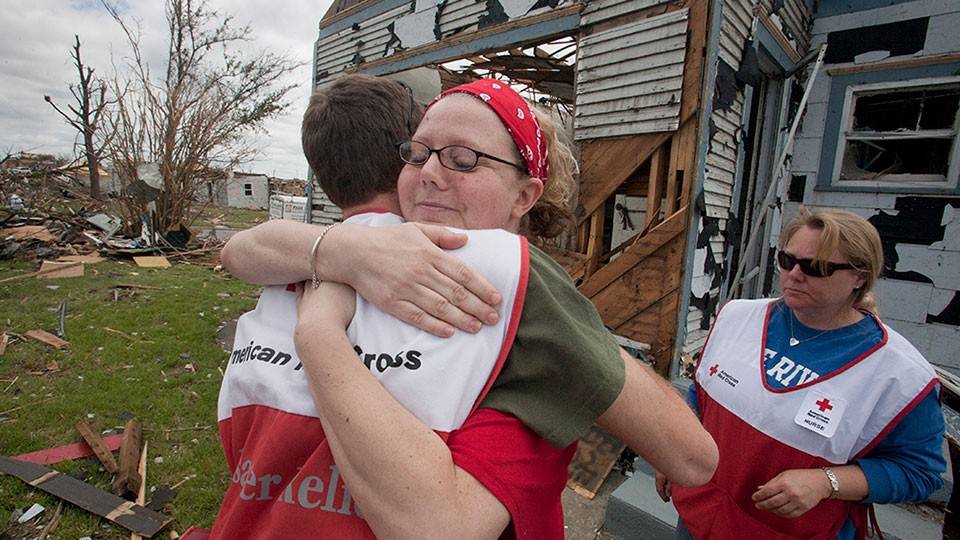 Two relief workers in Red Cross vests hug amid tornado debris. A damaged house is visible, conveying a sense of empathy and support.