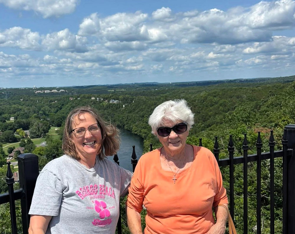 June St. Laurent and her daughter, Denise standing next to each other with a lush landscape and scattered clouds in the sky in the background.