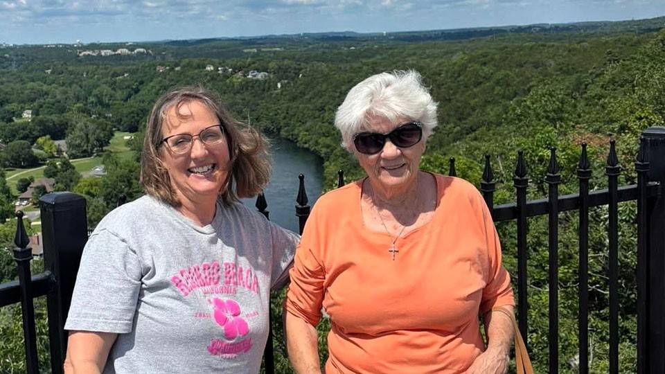 June St. Laurent and her daughter, Denise standing next to each other with a lush landscape and scattered clouds in the sky in the background.