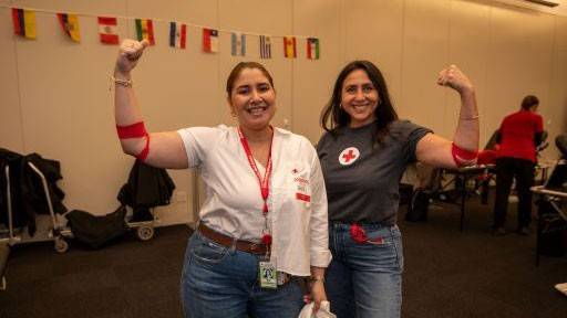 Two women smiling and flexing their arms with red bands, standing in a room with international flags on the wall.