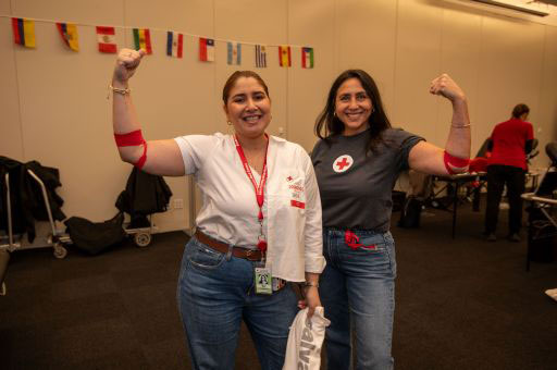 Two women smiling and flexing their arms with red bands, standing in a room with international flags on the wall.