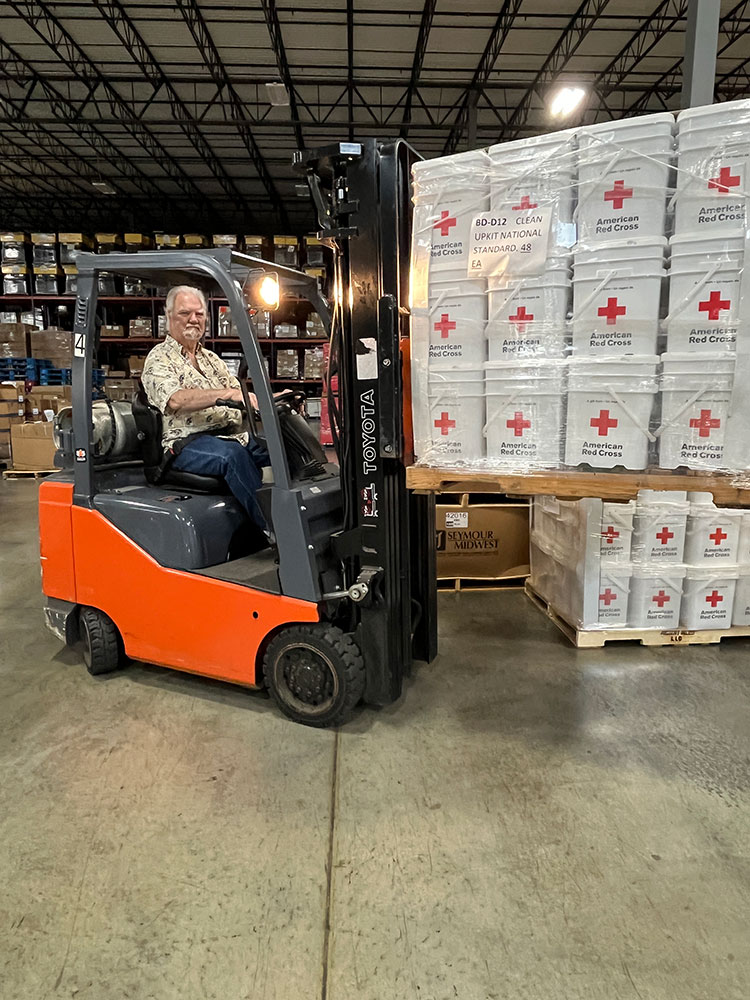 Richard Miller in a warehouse driving a forklift that has a load of Red Cross 5 gallon buckets.