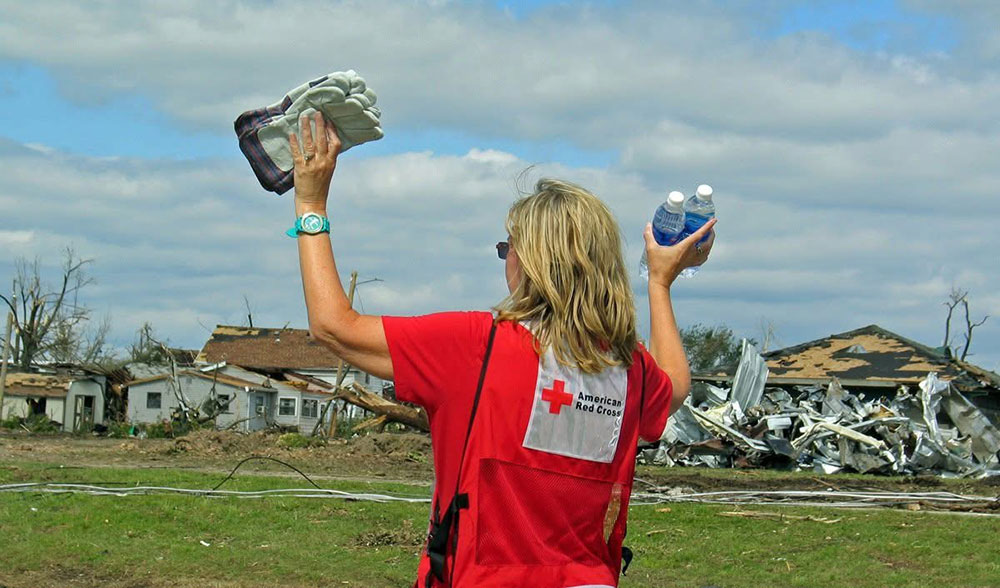 Sherri Odell wearing a Red Cross vest, holds up bottled water and work gloves next to houses that have been damaged.