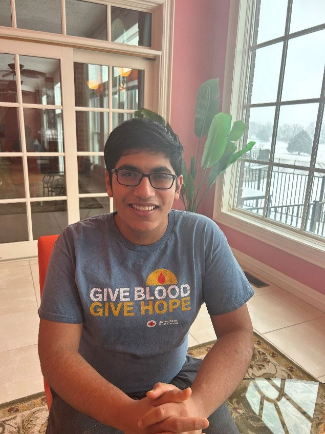 Shiv Bansal sitting in chair wearing glasses and a Red Cross Give Blood Give Hope shirt.