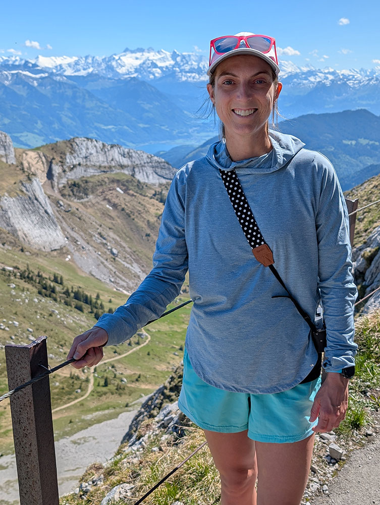 Amber Hanson smiling for picture while on a mountain next to a railing with snow capped covered mountains in the background.