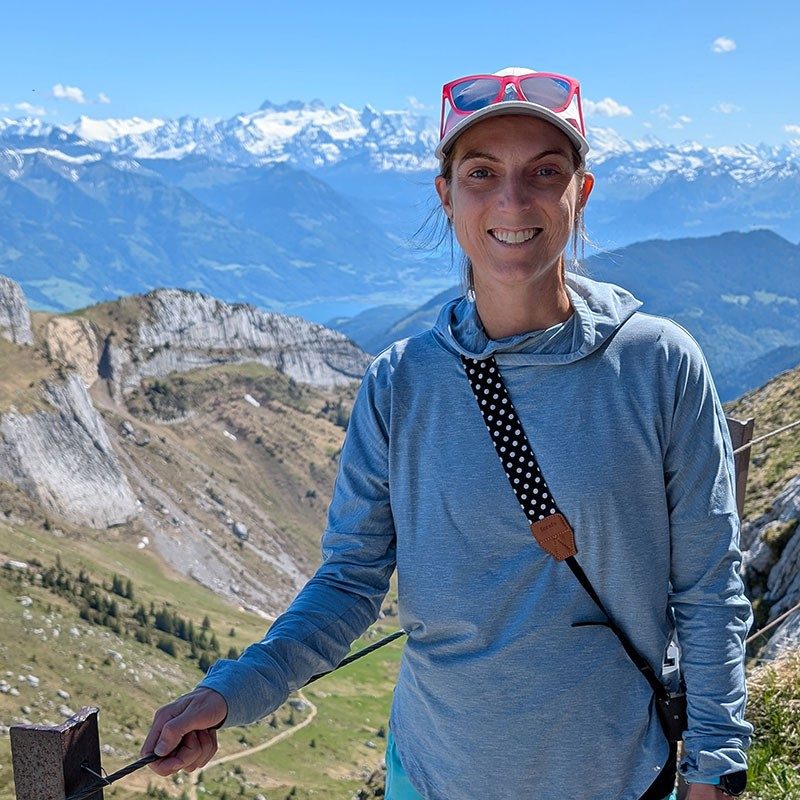Amber Hanson smiling for picture while on a mountain next to a railing with snow capped covered mountains in the background.