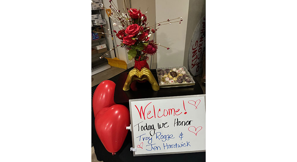 table with flowers in a vase, hands statue that's making a heart shape, a box of chocolates, heart shaped balloons, and a welcome sign.