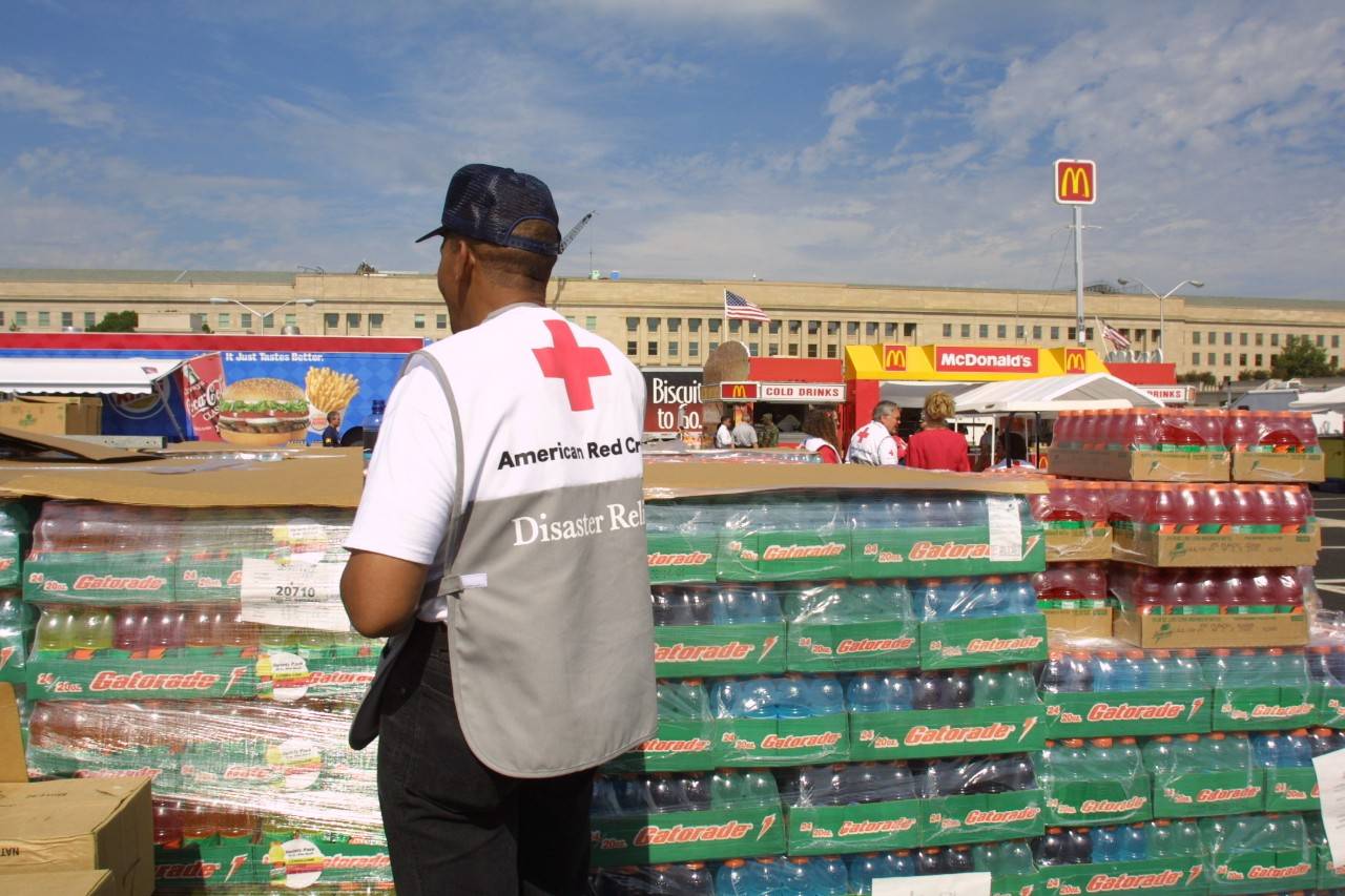Man wearing American Red Cross Disaster vest stands in front of a large number of cases of drinks in front of the Pentagon