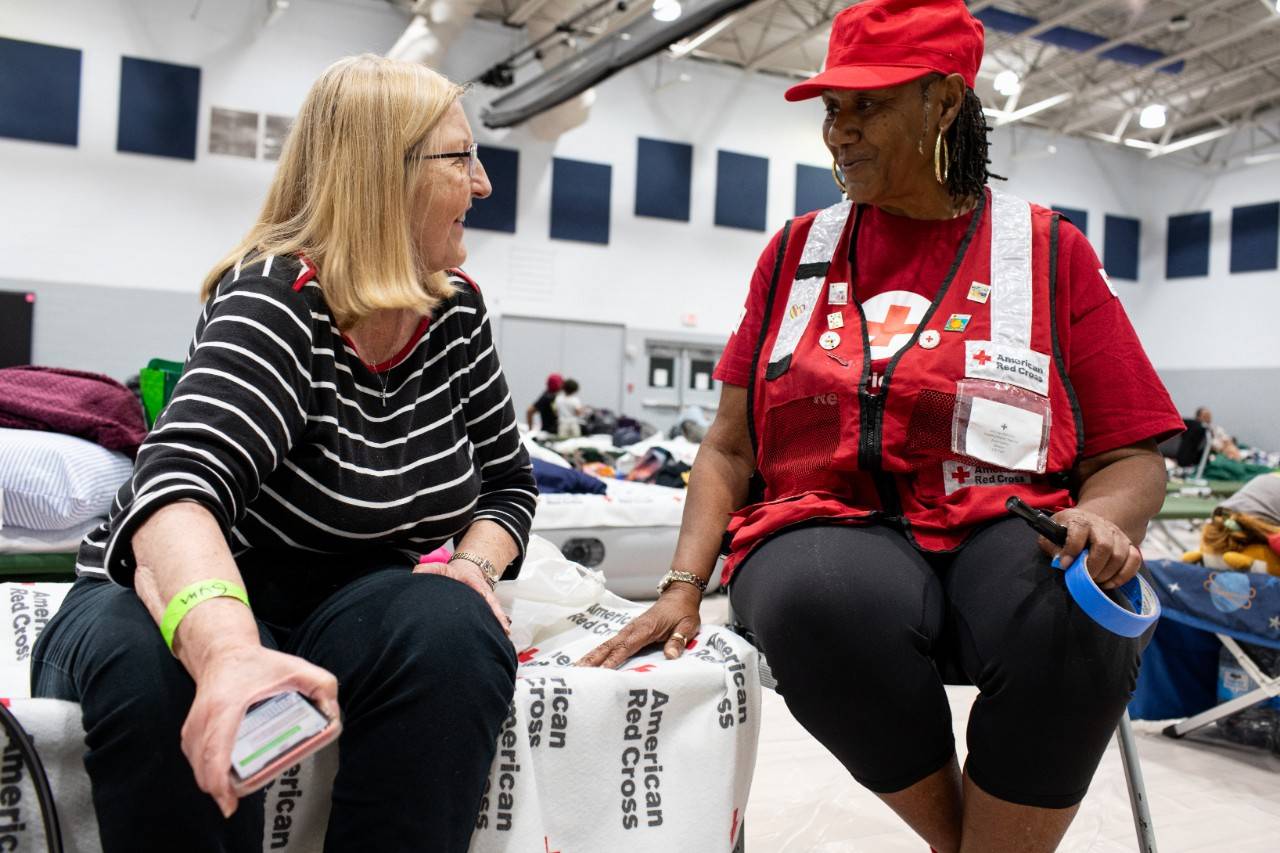 Volunteer Essie Hopkins sits and talks with a person who is living in a Red Cross shelter after Hurricane Milton. They are in a large open room full of cots. Essie is wearing a Red Cross vest.