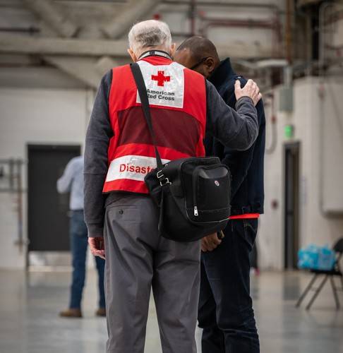A man wearing a Red Cross vest comforts a man and has his hand on the other man’s shoulder.