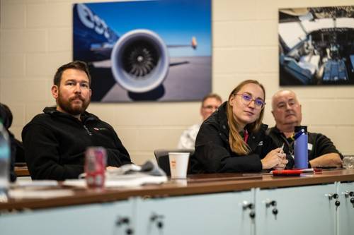 Two men wearing black shirts, one of which has the American Red Cross emblem, sits at a desk with a woman who is also wearing black, with two large pictures on the wall behind them, one of which shows a jet engine on the wing of a commercial airplane and the other showing the cockpit of an airplane.
