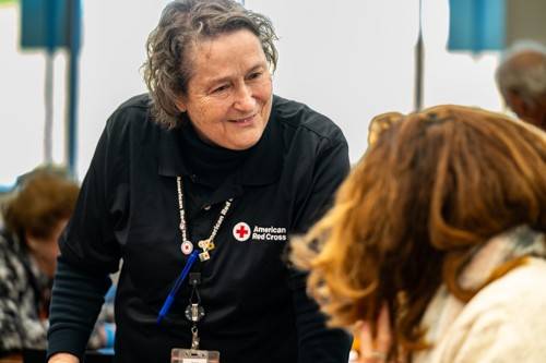A woman wearing a black jacket with the American Red Cross emblem talks to a woman while leaning forward on something.