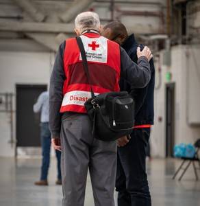 A man wearing a Red Cross vest comforts a man and has his hand on the other man’s shoulder.