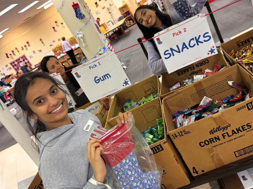 Young volunteers smile while sorting treats in a festive room. Boxes labeled "Gum" and "Snacks" are filled with candy, conveying a cheerful, holiday mood.