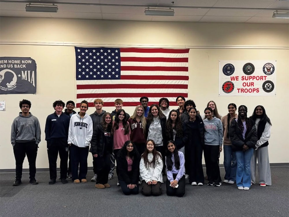 A diverse group of young adults stands smiling in front of an American flag and military support banners.
