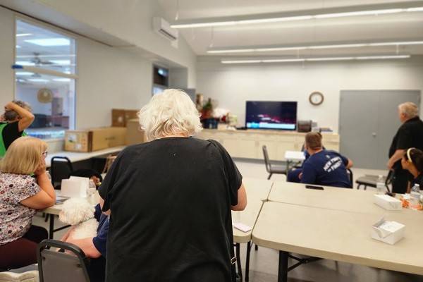 People in a room with folding tables and watching a tv that's on a wall.