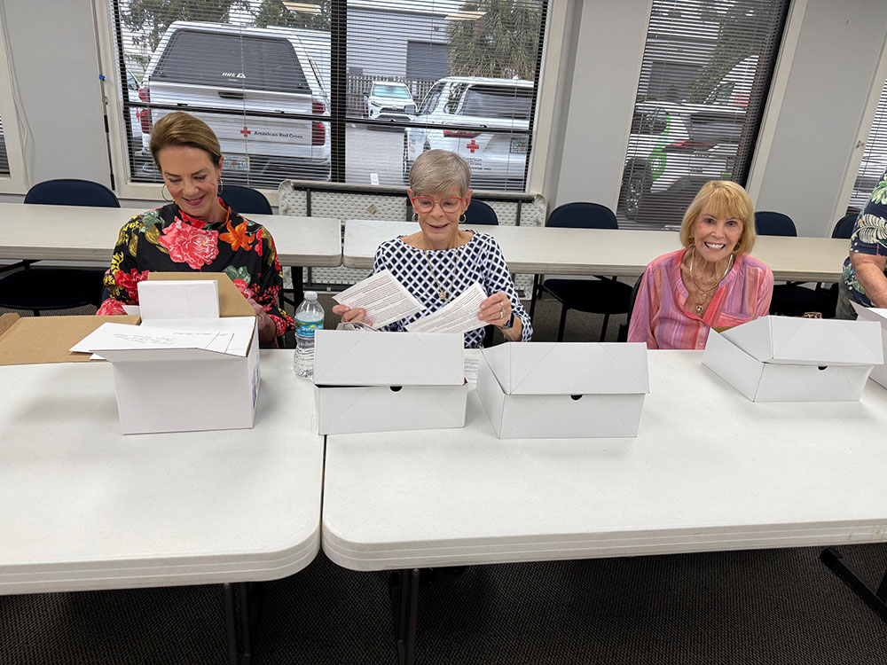 Three Red Cross staff members sitting at folding tables with boxes of paper on the tables.