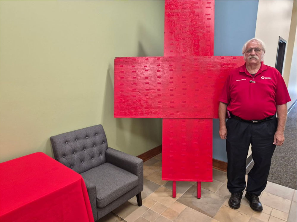 Michael Blain wearing a Red Cross shirt and standing next to a large Red Cross sign inside a room.