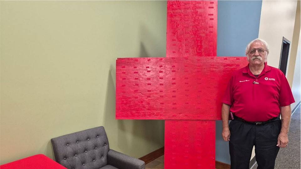 Michael Blain wearing a Red Cross shirt and standing next to a large Red Cross sign inside a room.