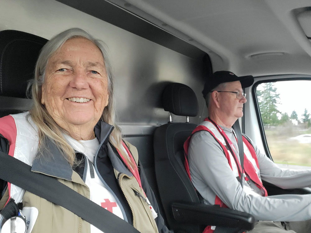Two people, wearing Red Cross vests, sit in a vehicle. The woman in the passenger seat smiles warmly. The man drives, looking focused.