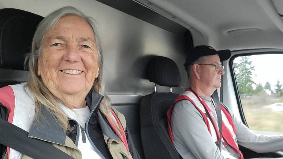 Two people, wearing Red Cross vests, sit in a vehicle. The woman in the passenger seat smiles warmly. The man drives, looking focused.