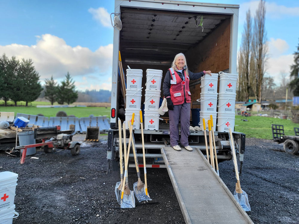 Nancy Diles in a Red Cross vest stands in a truck filled with Red Cross supplies. Shovels are propped against the truck amidst a rural, cloudy landscape.