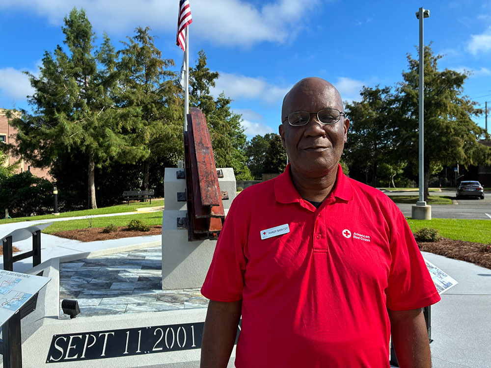 Randy Barkley wearing a Red Cross shirt and standing next to a 9/11 Memorial with a section of steel from the World Trade Center.