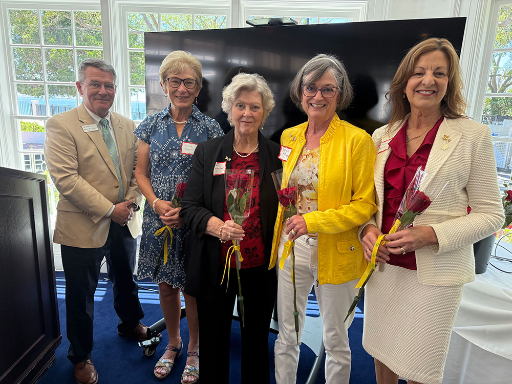 A group of five people smile indoors, four women and one man. Each woman holds a red rose.