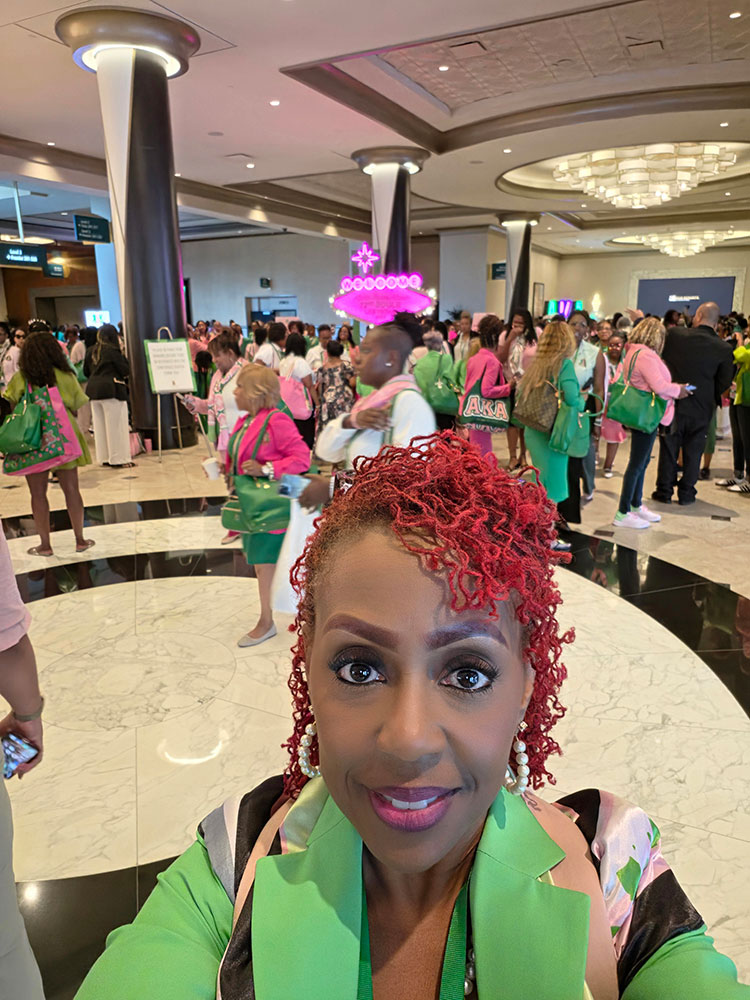 Dr. Andrea Pullen selfie wearing a green blazer with lots of people in the background in a room with marble floors.