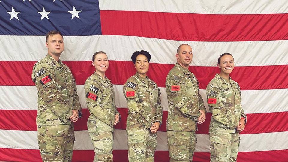 Angelica Sowynn and four others in military camo with a large American flag behind them.