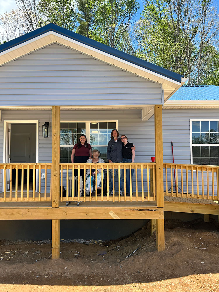A smiling group of four stands on a wooden porch of a light blue house with a sloped roof. A ramp is visible, indicating accessibility. Surrounded by trees.
