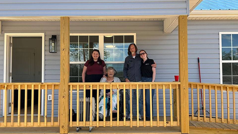 A smiling group of four stands on a wooden porch of a light blue house with a sloped roof. A ramp is visible, indicating accessibility. Surrounded by trees.