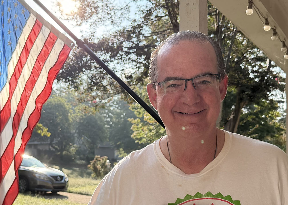 Frank Castillo wearing glasses and smiling with an American Flag in the background.