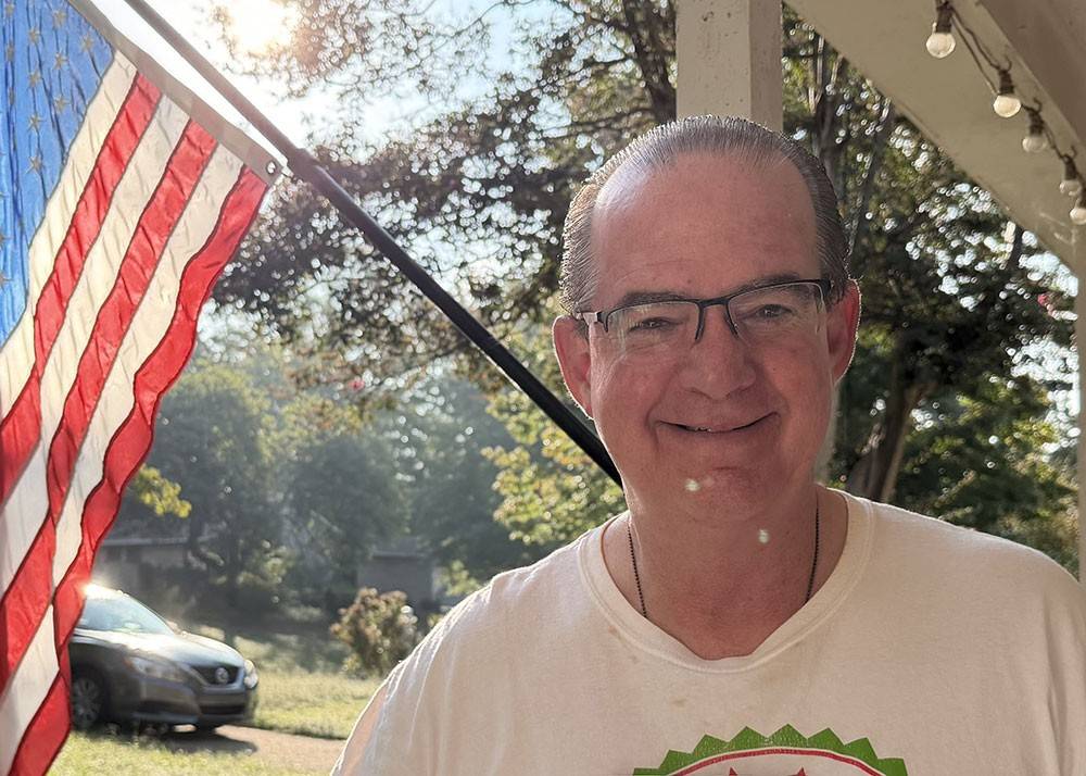 Frank Castillo wearing glasses and smiling with an American Flag in the background.