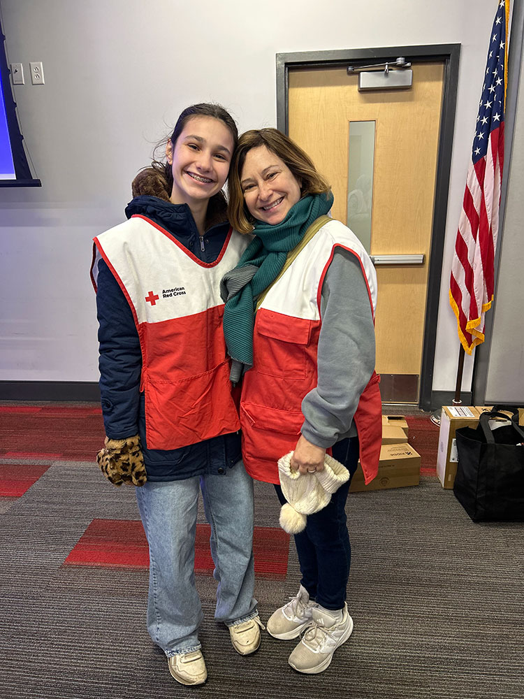Red Cross volunteers Lilah and Colleen Gross in their Red Cross vests with an American flag in the background.