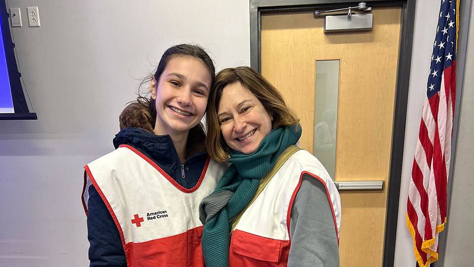 Red Cross volunteers Lilah and Colleen Gross in their Red Cross vests with an American flag in the background.