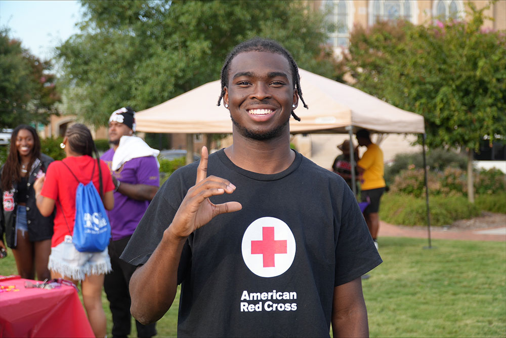 Hunter Howard smiling in an "American Red Cross" shirt gestures with one hand at an outdoor event with a tent and other people in the background.