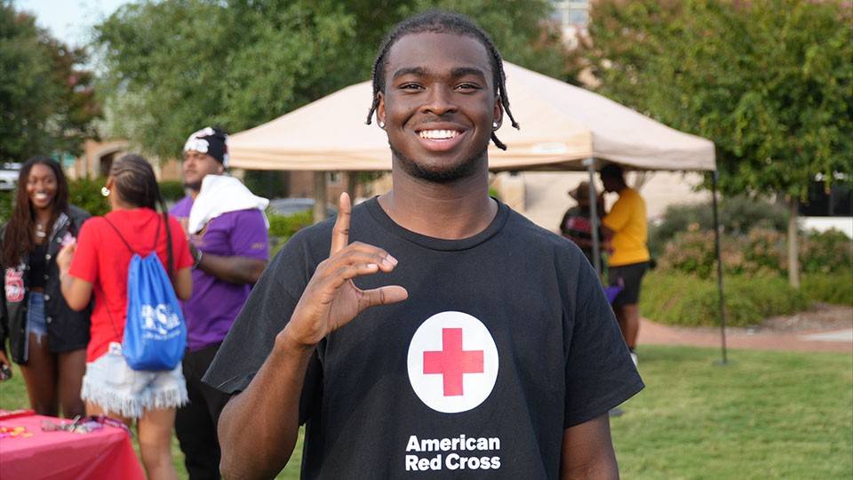 Hunter Howard smiling in an "American Red Cross" shirt gestures with one hand at an outdoor event with a tent and other people in the background.