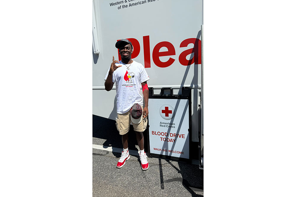 Hunter Howard smiling stands in front of a Red Cross blood drive bus, wearing a white T-shirt and red-accented shoes. A sign reads "BLOOD DRIVE TODAY."